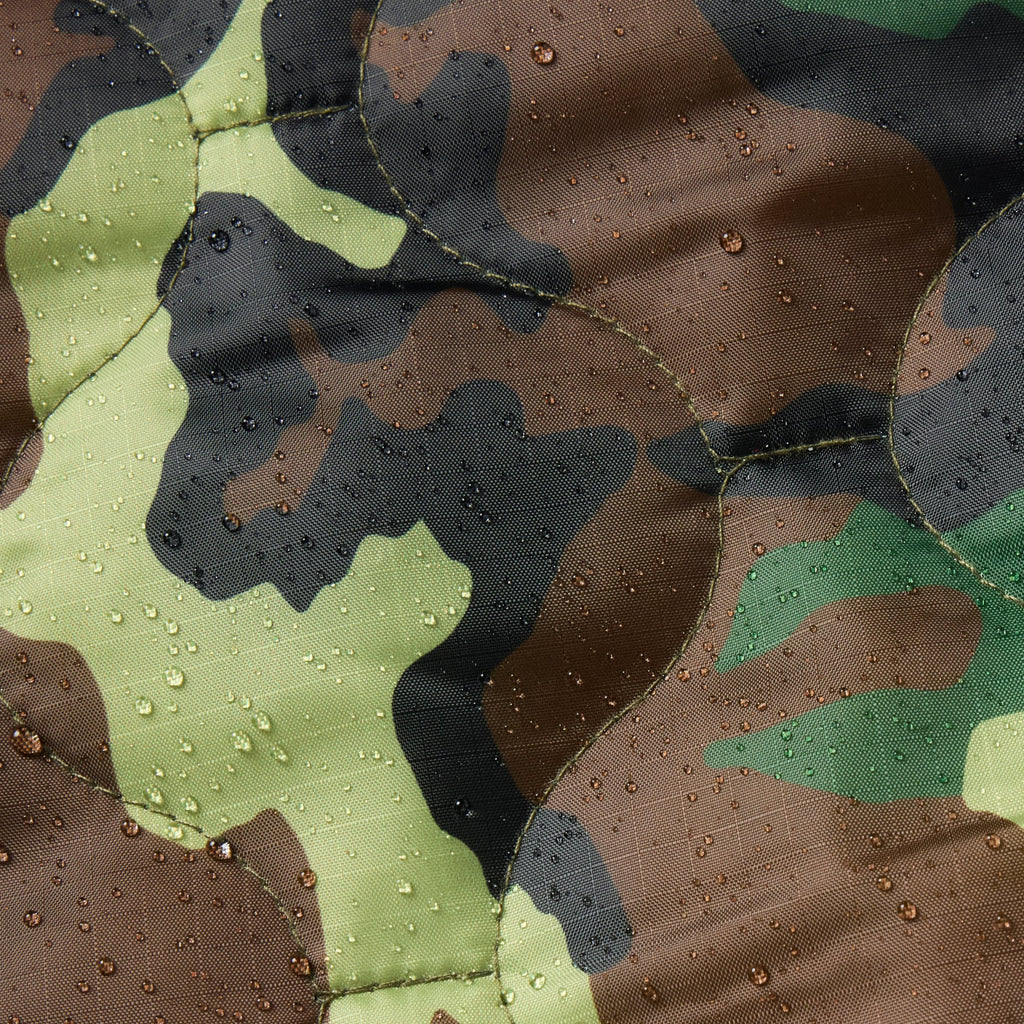 Close-up of a quilted fabric with a woodland camouflage pattern in green, brown, black, and tan, covered with water droplets.